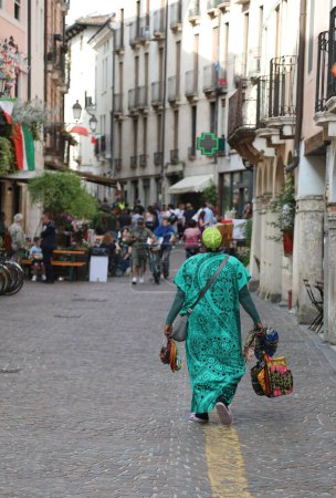 Frau mit grünem Tuch verkauft Glücksketten und Glücksbringer an Passanten in einer historischen Straße im europäischen Stadtzentrum
