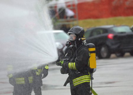 Photo for Firefighters with oxygen tanks and protective helmets spray a powerful stream of foam to extinguish the fire while it rains - Royalty Free Image