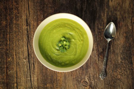 Top view of a broccoli soup in a white bowl with a spoon on a rustic wooden background