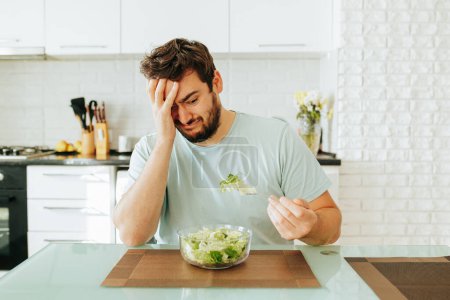 Photo for A sad young man looks longingly at a salad, holds a fork with greens in his hand, and a plate of salad in front of him. An exhausting diet haunts, hates healthy food. Stop diet. - Royalty Free Image