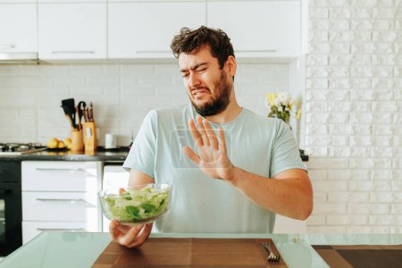 A young man, grimacing, looks towards the salad, how you don t want to eat it. Located in the kitchen. Filming inside. Stop diet. Front view.