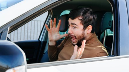 Photo for A young man looks in the side mirror of his car, let go of the steering wheel and screams. Probably saw something on the road and was scared or surprised. Peoples reactions to different situations. - Royalty Free Image
