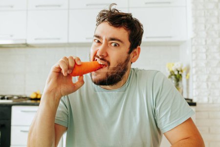 Frontansicht eines jungen Mannes, der mit grimmigem und unglücklichem Gesichtsausdruck in eine rohe Karotte beißt. Gesunde Ernährung ist ein Muss, um gesund zu sein. Schluss mit Diät, hallo gute Laune.
