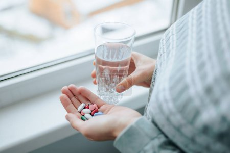 Unrecognizable top view woman with full palm of pills and glass of water stands by window. Girl must take medication and is preparing to do it. Concept of health care. Pharmacological preparations.