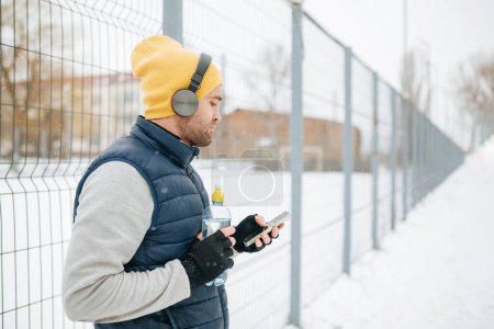 Foto de A young guy takes a break from his wintertime run to hydrate himself with a drink of water. The solitary runner is caught in action, using his smartphone to listen to music while keeping - Imagen libre de derechos