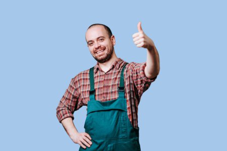 Foto de Joyful farmer with thumbs up and looking at camera, in studio Contented farmer in blue studio shot, Ebullient engineer with beard in overalls. copy space and blue background - Imagen libre de derechos