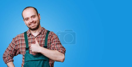Foto de A proud, excited young farmer with a beard is seen in this studio photograph, giving a thumb up and dressed in overalls. A contented, enthusiastic young farmer, wearing coverall copy space - Imagen libre de derechos