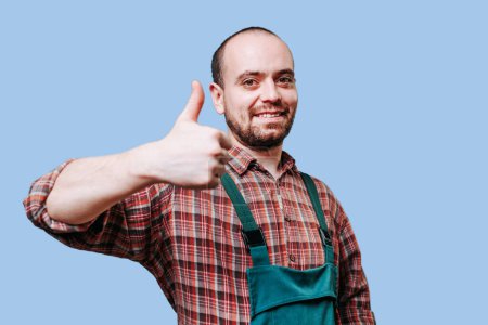Foto de A portrait of a nice, smiling, happy, bearded young farmer workman with thumb up looking at the camera, wearing overalls, and standing in a studio shot. copy space, blue background - Imagen libre de derechos