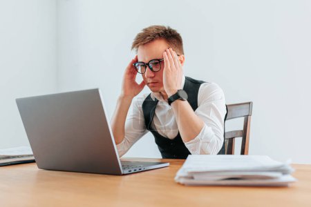 Photo for A guy in eyeglasses looks exhausted and overwhelmed as he works at his computer, struggling to manage the stress and workload of his job. Eyestrain and Overload The Strain of the Office Job - Royalty Free Image