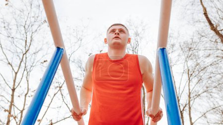 Photo for Outdoors and Athletic A Young Sportsmans Gymnastics Practice. A young athletic man in a red t-shirt trains his muscles on the parallel bars outside in the fresh air. - Royalty Free Image