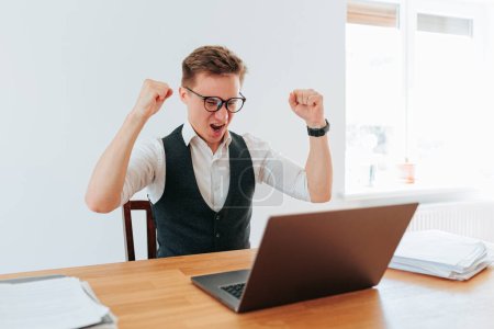 An indoor shot shows an office worker sitting alone at his desk, hands raised in victory after completing a challenging assignment. The satisfaction and happiness he feels is a testament to his skill