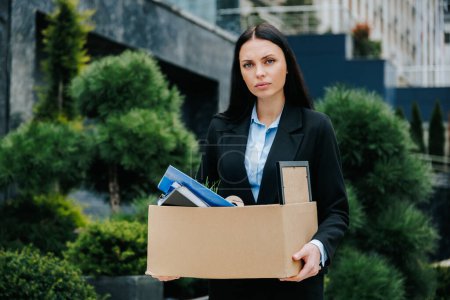 An image of a woman sitting outside, looking sad and unemployed after losing her job. The Reality of Unemployment Woman Holding Box in Empty Parking Lot