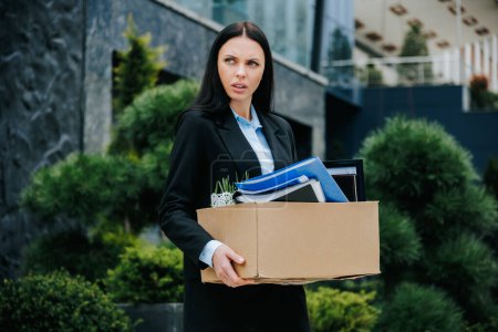 Téléchargez les photos : Un employé de bureau debout à l'extérieur avec une boîte, dépeignant la perte de son emploi et de son travail. Les difficultés de la femme sans emploi tenant boîte après le licenciement - en image libre de droit