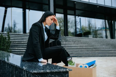 Une femme est montrée assise sur une boîte en carton, tenant un panneau indiquant qu'elle a perdu son emploi et qu'elle a l'air bouleversée et en détresse..