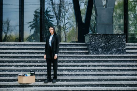 Foto de Una mujer está de pie en las escaleras de un edificio de gran altura con una caja de cartón, lo que refleja la realidad de la pérdida de empleo y el desempleo durante una época de crisis y recesión económica. - Imagen libre de derechos