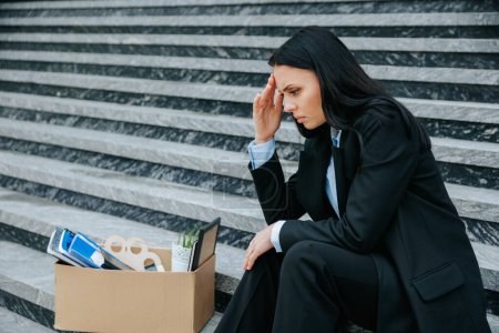 Foto de Una foto poderosa de una mujer sentada en las escaleras, sosteniendo una caja de cartón, que representa la pérdida física y emocional de su trabajo despedido y su falta de trabajo. - Imagen libre de derechos
