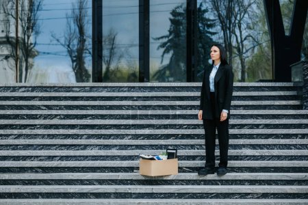 Foto de Una joven mujer está de pie en las escaleras de un edificio urbano con una caja de cartón, mirando hacia abajo y preocupada por perder su trabajo y enfrentarse al desempleo. Desempleados en la escalera de la ciudad con caja de cartón - Imagen libre de derechos