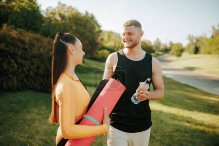 Photo for Two young adults, wearing fitness attire, are outside engaged in conversation and winding down after their workout. The man carries a water bottle, and the woman has a fitness mat - Royalty Free Image