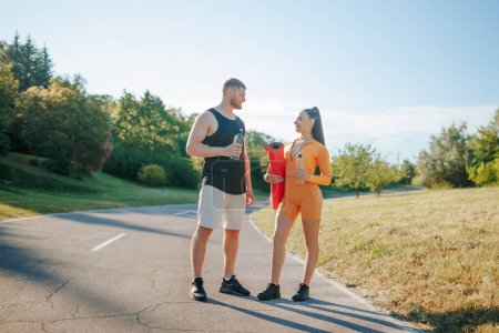 Photo for Two young adults, both athletes, a man, and a woman, in sportswear, engaging in conversation and enjoying the outdoors after their workout. while the woman is holding a fitness mat. - Royalty Free Image