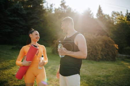 Photo for Two athletes, a man and woman, dressed in sportswear, enjoying each others company outdoors after their training. The man holds a water bottle, and the woman carries a fitness mat. - Royalty Free Image