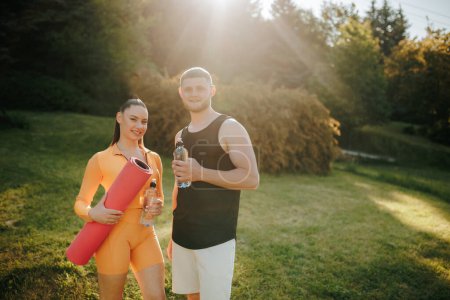 Photo for A man and woman, dressed in fitness attire, are outside, talking and winding down together after their exercise routine. The man carries a water bottle, and the woman holds a fitness mat. - Royalty Free Image