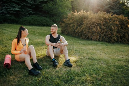 Photo for Two athletes, a man and woman, wearing sportswear, enjoying each others company outside after their workout. The man holds a water bottle, and the woman is holding a fitness mat. - Royalty Free Image