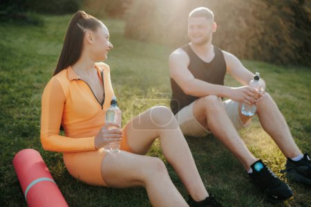 Photo for Two athletes, a male, and a female, dressed in fitness clothing, chatting and taking a break outside after their exercise routine. The man has a water bottle, and the woman holds a fitness mat. - Royalty Free Image