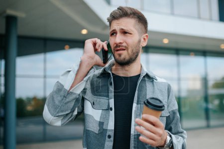 Ein ernst dreinblickender junger bärtiger Mann diskutiert draußen auf seinem Smartphone. Junge Erwachsene mit Handy drücken Frust während eines Gesprächs aus