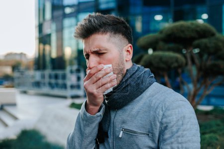 Erleben Sie die Qualen eines jungen bärtigen hispanischen amerikanischen Mannes, der unter Grippesymptomen leidet, erbarmungslos niest und hustet, dargestellt in einem Straßenbild der Stadt.