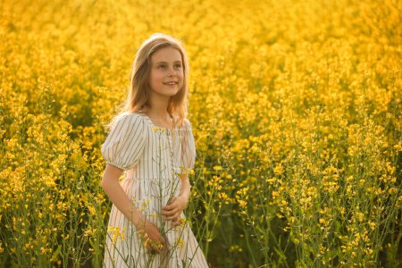 Téléchargez les photos : Portrait d'une belle petite fille heureuse dans la robe, sur un fond jaune naturel avec du colza fleuri. rustique, espace de copie - en image libre de droit
