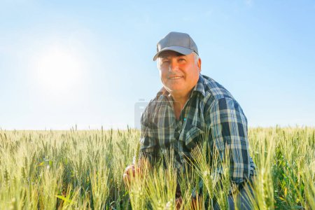 Téléchargez les photos : Elderly male farmer in cap looking into distance with pensive face while sitting on haunches in agricultural field on summer day. Mature worker sitting in field - en image libre de droit