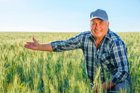 Joyful elderly male worker in cap looking at camera and pointing away while sitting in field with green plants in countryside. Smiling farmer pointing away in field