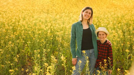 Portrait young mother woman stands in yellow flowering rapeseed embrace little daughter girl in hat looking at camera smiling with kid enjoying together outdoors in nature