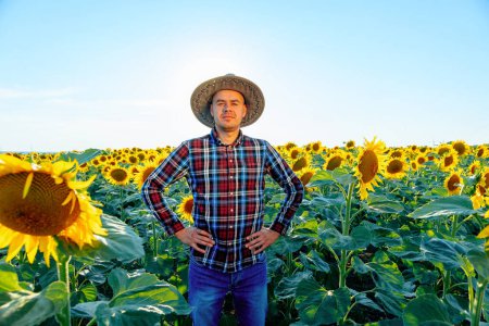 Ein junger Bauer hält seine Arme auf den Hüften und lächelt, während er inmitten einer Sonnenblume steht. Der Mann mag sein schickes Feld. Mensch und Kopierraum.