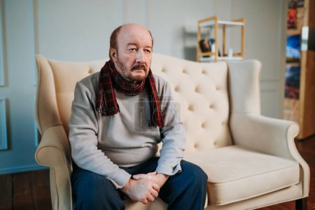 Aged man with a sad look sits on the sofa and looks somewhere into distance. Alzheimer struck a man at a venerable age. Distant look, no thoughts, scarf around his neck, a doomed look. Front view.