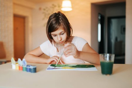 Das kleine Mädchen sitzt am Tisch und zeichnet ein Bild in das Album, während es sie ansieht. Beide Hände auf dem Tisch, konzentriert schauen sie auf helle Kleidung. In der Nähe ein Glas Wasser und Farbe. Frontansicht.