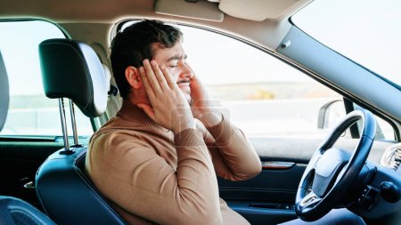 Side view male driver clutches his head with his hands and grimaces from the pain in his head. The guy cant keep going down the road because its hard for him. Headaches and emotions.