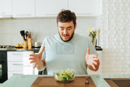A young man sits in the kitchen in front of a glass bowl of salad with mixed feelings. He looks at the salad with bulging eyes. Greens again