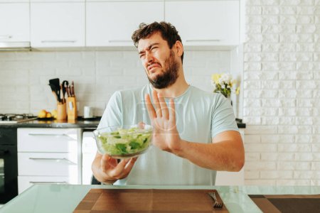 Closing eyes and turning his head to the side, a young man holds a bowl of healthy breakfast in his hand. I want to eat something else, but you need to follow a diet. Healthy food concept. Stop diet.