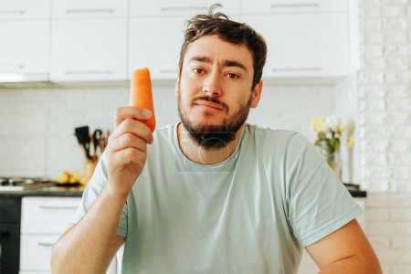 Front view looking at the camera a young man indifferently and slightly tormented holds a raw carrot in his hand. He must be on the right diet. This puts him in a bad mood.
