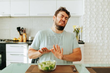 A nice young guy man sits in the kitchen in front of a bowl of salad and holds his hands in front of him. A person monitors his health and weight, hoping to quickly finish the diet. stop diet.