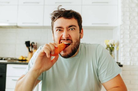 Front view looking at camera a young man makes a grimace and bites raw carrots with anger, does not like vegetables. Does not want to go on a meat free diet. Vegan food spoils the mood. Stop diet.