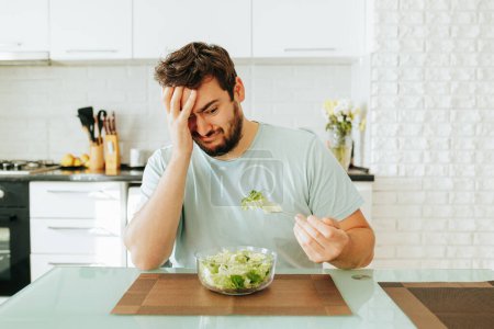 A young guy holds a fork with a green salad in his hands and looks doomed at this food. Fresh herbs in a bowl look appetizing, but that does not convince a young man to be in a good mood. Stop diet.