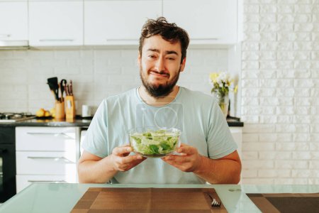 A young man with a martyred expression looking at the camera and holds a glass bowl of salad in his hands. The guy suffers from the need to eat vegetables and salad. Stop diet. Front view.