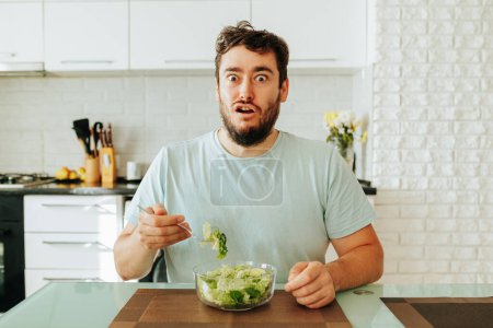bizarre Young man expression horror on face parted mouth opened eyes wide hold a fork with salad in hand. A man should eat salad to be healthy, but he is tired of such food. Stop diet.