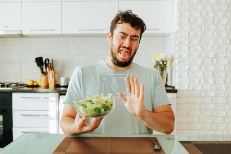 Sitting in a modern kitchen holding a bowl of fresh salad in hands, he grimaces and fences himself off from healthy food. Good eating habits are not always pleasant and tasty. Front view. Stop diet.