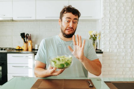 In one hand, a young man has a bowl of salad, the other shows a stop to salad and vegan food. Greens and vegetables are very healthy, but the guy is not tasty. Front view. Stop diet.