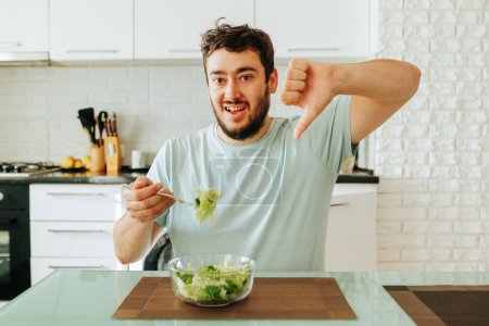 Sadly showing a dislike, a young guy holds a fork with a green juicy salad in his hand. The diet is useful, but it spoils the mood. Stop dieting. It is time to improve your mood.