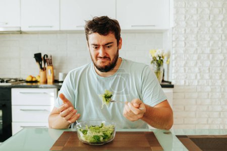 A young man shows the emotion of disgust in his hand holding a fork with a juicy green salad. A bowl of healthy dinner is on the table, but no one wants to eat it. Stop diet.