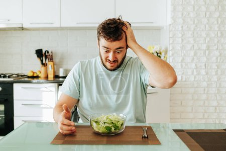 Ein panischer Blick auf einen grünen Salat bei einem jungen Mann auf Diät. Das alles will er nicht essen, muss es aber. Nochmals gutes Essen. Gesunde Ernährung ist langweilig. Schluss mit Diät.